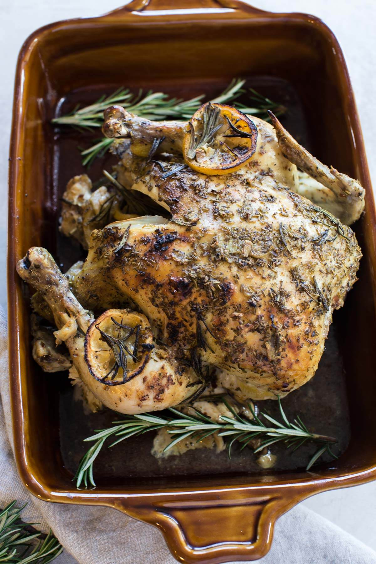 overhead shot of cooked whole chicken with rosemary sprigs, fresh herbs and lemon slices